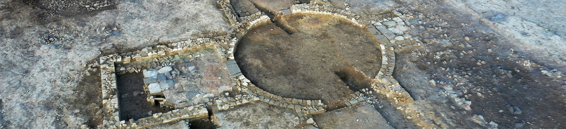 Low stone structures in excavated ground. The main structure includes a central circle of stone with three rectangular rooms radiating from it.