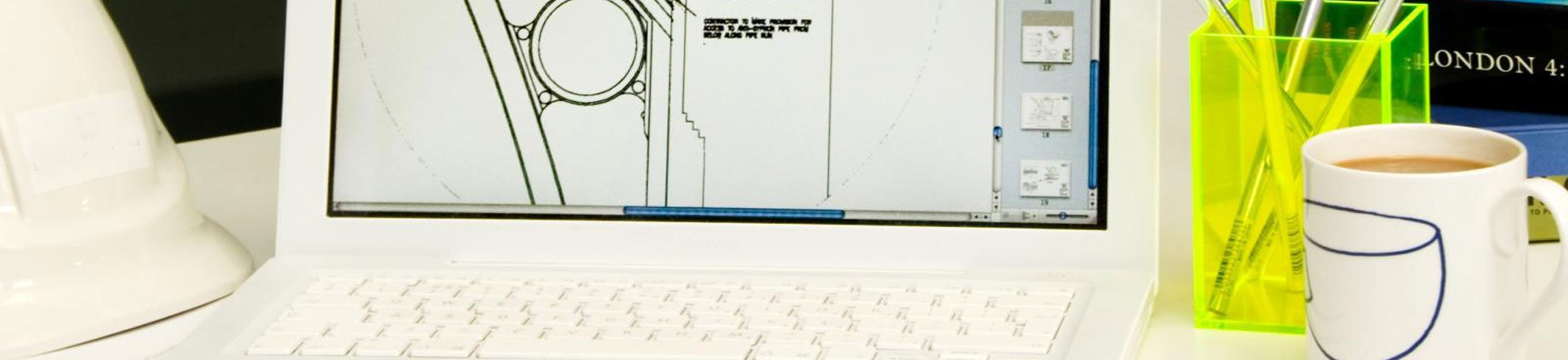 Detail of office environment including building plans on a laptop screen and a hard hat on the desk next to it.