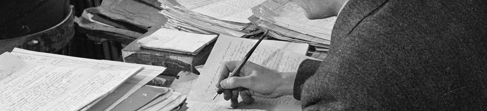 A man wearing a suit writing at desk surround by piles of paper