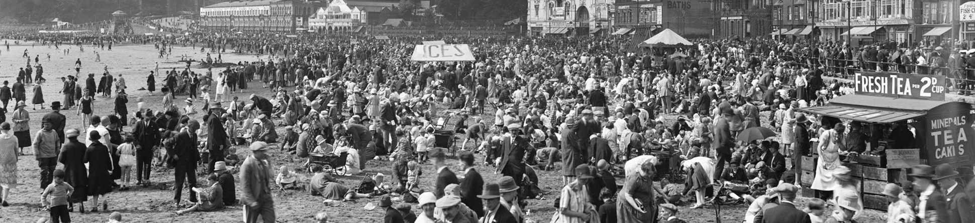 An archive photograph of a seaside resort beach scene with crowds in 1920s clothing.