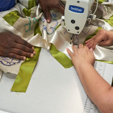 Two pairs of hands work together to sew together a ribbon on a panel.