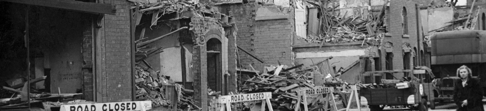 In front of the damaged houses are 'road closed' signs and a woman walking along in the road.