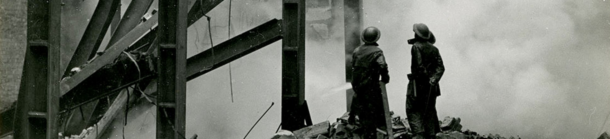 Two fire fighters stand on a pile of rubble next to steel girders looking up at a bomb damaged building partially shrouded by smoke.