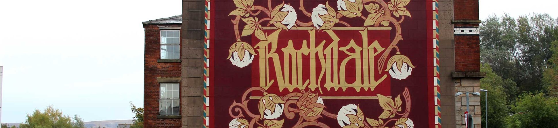 A mural featuring the name of the town, its Latin motto, cotton bolls and red roses, created on the side of a mill building.