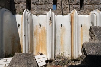 A row of stained and damaged urinals standing in the open air, in front of a stone wall. In the foreground sections of former wall tiles are lying on the ground in front of the urinals.