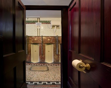 A colour photo showing the view within a dark wood toilet cubicle, looking out across a decoratively tiled floor towards a row of granite and porcelain urinals.
