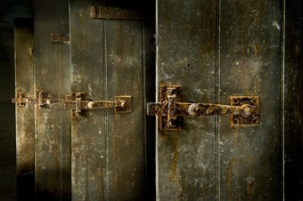 A detail view showing the rusty, wrought iron latches on the wooden plank doors of a row of toilet cubicles.