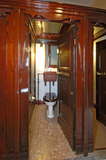 A colour photograph showing the view looking through the open door of a toilet cubicle. The door bears the word ‘GENTLEMEN’ and stands within an ornately shaped doorway. The cubicle contains a toilet pan with ‘CRAPPERS’ cistern. Adjacent is a partially visible Ladies cubicle.