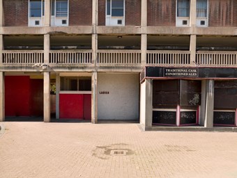 The ground floor of a block of flats on a mid-20th century council housing estate. In the centre of the image is the entrance to the ‘Ladies’. Above the windows of a former pub a sign reads ‘Traditional Cask Conditioned Ales’.