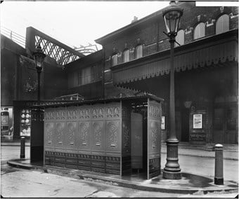A black and white photo showing the exterior of a rectangular cast iron urinal building with gas lamp posts at either end. Brixton railway station in the background and a viaduct partially visible to the left.