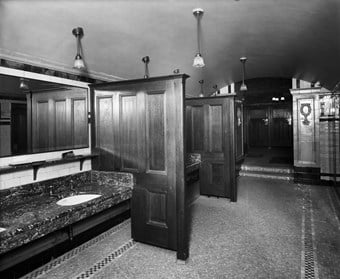 The interior of ladies lavatories showing the wash basins in the foreground. The basins, which are mounted in marble worktops, are separated by dark wooden screens allowing privacy for users. Large mirrors hang on the wall in front of the basins. The floor is finished in mosaic tiles and the walls are also decoratively tiled. Electric light fittings hang from the ceiling above the basins.