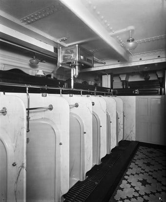 A row of urinals beneath a low, riveted ceiling on board ship. Each urinal has a metal bar extending across it, presumably to aid with balance on rough sea crossings.