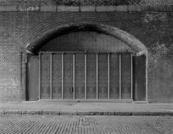 A black and white photograph looking across a cobbled street towards the brick arch of a viaduct. A decorated cast iron encloses the interior of arch, behind which are the urinals.