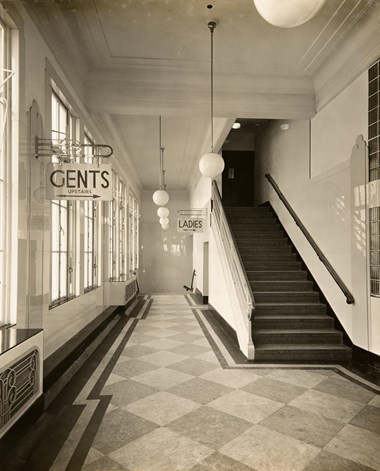 An interior view looking along a chequerboard carpeted landing with a flight of stairs to the right of the image. On the wall in the left foreground is a sign for the ‘GENTS’, which is located upstairs. Further along the corridor is the sign for the ‘LADIES’.