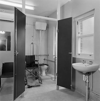 The interior of the toilet facilities looking towards the open door of a cubicle inside which is a toilet with an empty wheelchair to the side.