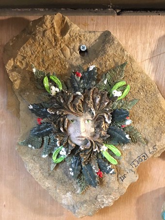 A stone roof tile or shingle painted with a "green man" style motif by a 21st-century artist.