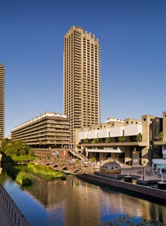 Colour photo showing the Barbican Centre next to a narrow lake, with a high tower in the background.