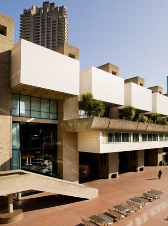A colour photo showing a side view of part of the Barbican complex. A rectangular sand coloured apartment building with small palm trees on balconies can be seen in the foreground, with a tower block in the background.