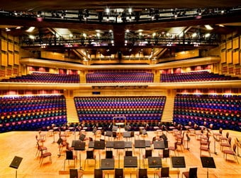 A colour photo showing the interior of a concert hall in the Barbican centre. Rainbow coloured rows of seats extend in the background, overlooking the stage in the foreground with seats for musicians.
