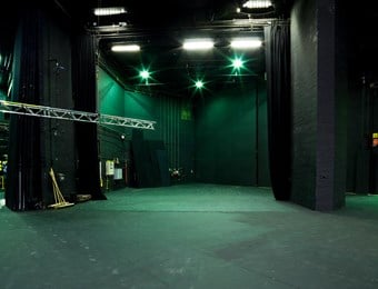 A digital colour photograph showing the backstage area of the Pit theatre at the Barbican centre. Green lights shine on the dark grey walls, with various equipment stored in the background.