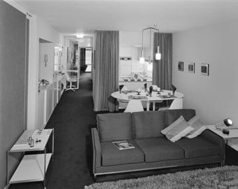A black and white photo showing the interior of a flat with a large sofa and sitting room in the foreground, and a kitchen area in the background.