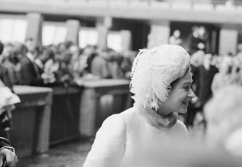 A black and white photo showing Queen Elizabeth II dressed in white at the Barbican Arts Centre, with a blurred view of a crowd of people in the background.