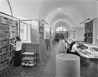 A black and white photo showing the interior of the Guildhall School of Music and Drama Library. The library’s floors and walls are partially covered with bricks, and three people are reading books in the area.