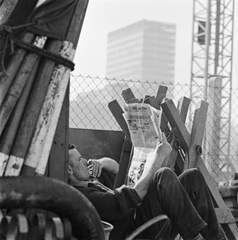 A black and white photo of a man sitting down with a newspaper, smoking a cigarette. The man is surrounded by the Barbican building site in progress, with a crane and tower in the background.