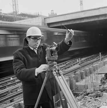 A black and white photo showing a surveyor wearing a hard-cap, taking measurements with a theodolite instrument, in front of a railway line with a speeding train passing.