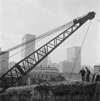 A black and white photo showing a building site with a crane in the background, holding a bucket swinging in the middle of the photo. Tower blocks are visible in the background and a few people are on site.