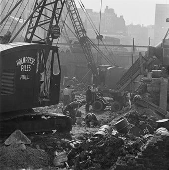 A black and white photo showing the building site of the Barbican estate, with a crane in the foreground that has a sign on it saying ‘Holmpress Piles of Hull’. Men are working in the background amongst bricks and debris.