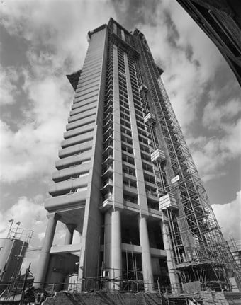 A black and white photo showing the Cromwell Tower under construction. The tower is seen from a low angle, with scaffolding surrounding it.