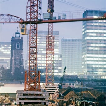 A colour photo showing the Barbican estate under construction, with a red crane in the foreground. St Giles Church and modern tower blocks can be seen in the background.