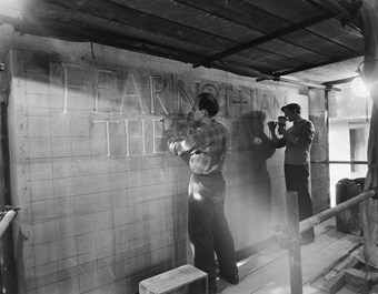Two stonemasons engrave a large stone wall.