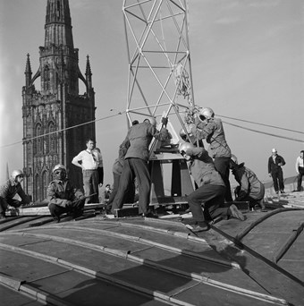 Men in helmets working to manoeuvre a metal frame. Six other men stand observing. The spire of the old cathedral is in the background.
