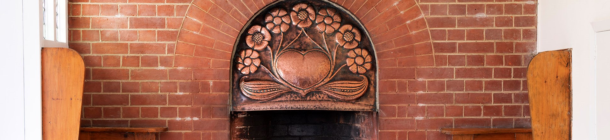 Interior of dining room with the ingle nook on its western wall having fixed seats at either side with cut-out heart shapes. The hearth has a semi-circular opening and a beaten copper fire hood. The copper fire hood has a central heart surrounded by flowers and leaves, emblematic of the Immaculate Heart of Mary.