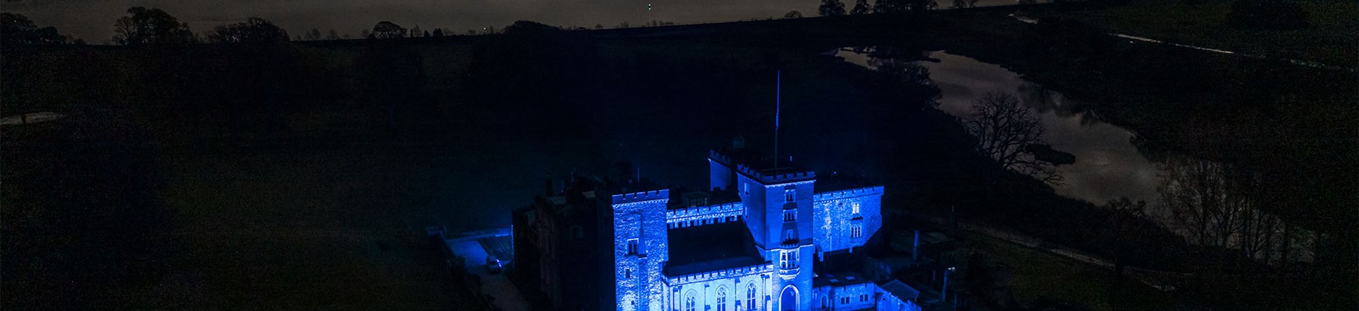 Night-time image showing Powderham Castle lit up blue for key workers against a cloudy sky and the lights of a town reflected in the river.