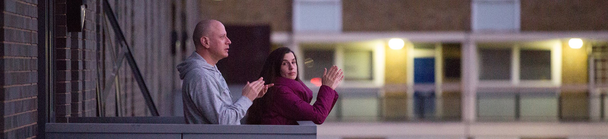 Man and woman standing on the balcony of a block of flats clapping their hands