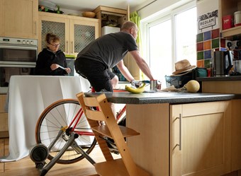 A man rides an indoor bike in his kitchen beside a woman holding a measuring tape above an ironing board.