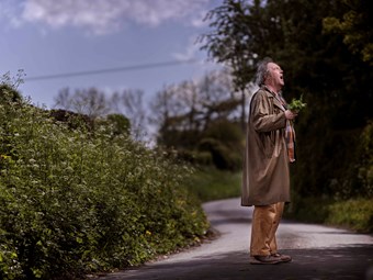 An elderly man stands in an empty country lane. He is holding a bunch of wildflowers, gasping up at the sky.