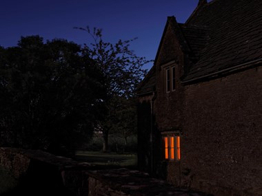 Orange light shines through a window at twilight. The home is made of Cotswold stone.