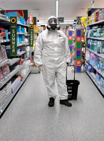 A man in full HAZ mask and suit carries a wheelie basket down a supermarket aisle.