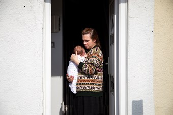 A woman stands at an open front door holding a newborn baby. She wears a brown fair-isle cardigan. The baby is in a white romper.