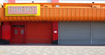 A large store front with corrugated orange facade and red shutters is closed. The shop is called 'sunspot; these words are signposted in pink and yellow. It is a warm sunny day.