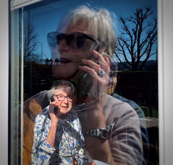 A woman has a conversation with her elderly mother via mobile phone. They can see each other through a sunny window.