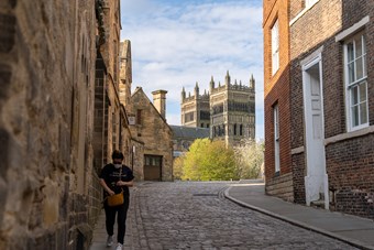 A woman in sunglasses and a face mask rummages inside a brown handbag whilst walking down an otherwise empty street in Durham. There are church towers in the background and a cobbled lane in the foreground.