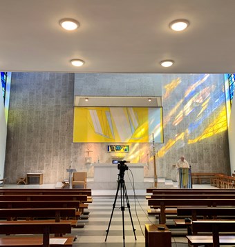 A priest gives a sermon to a video camera in an empty church. Light streams in from a yellow stained glass window.
