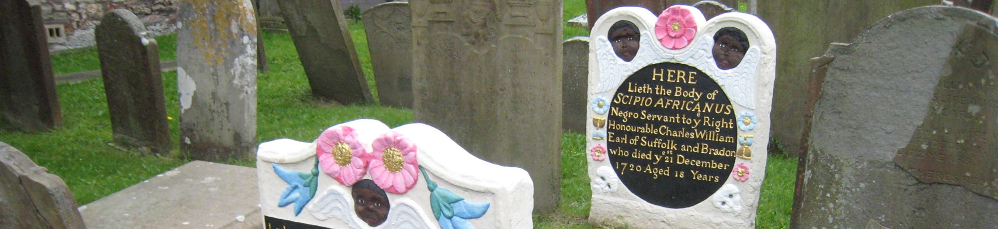 Headstone and footstone, with shaped tops. The freshly painted memorial stands out amongst unpainted stones. Carving includes winged Black cherubs, and flowers. The headstone carries the details of the subject’s life; the footstone carries a verse.