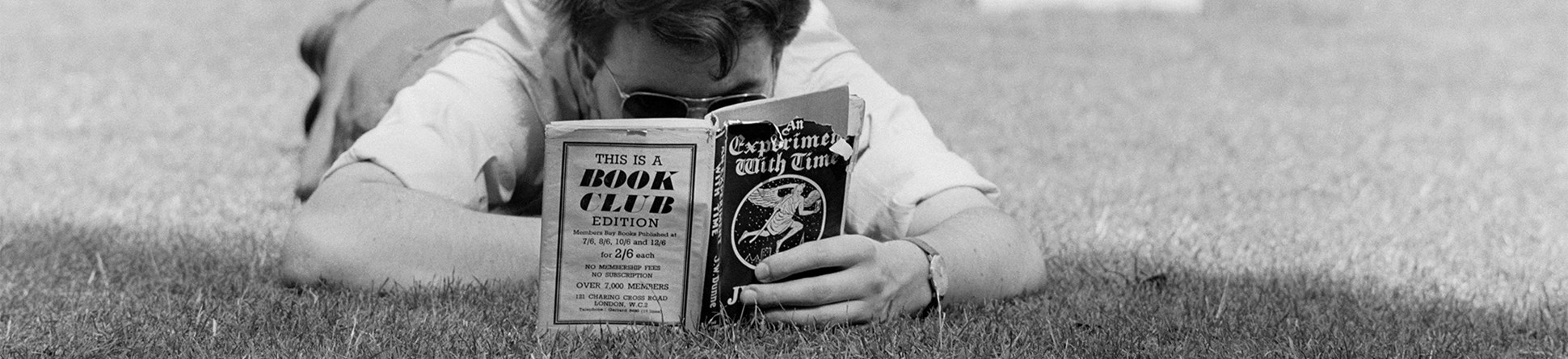 Black and white image of a man viewed head-on lying flat on the grass, engrossed in a book.