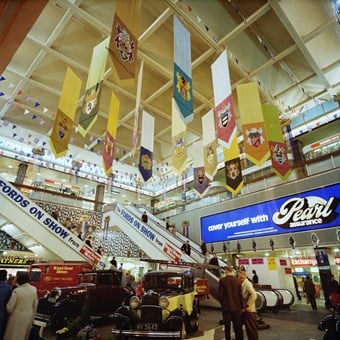 Colour photograph showing the Centre Court of the Bull Ring shopping centre. Vintage cars are on display as part of the ‘Fords on Show’ exhibition from Bristol Street Motors, and flags are up for the Queen’s Silver Jubilee.